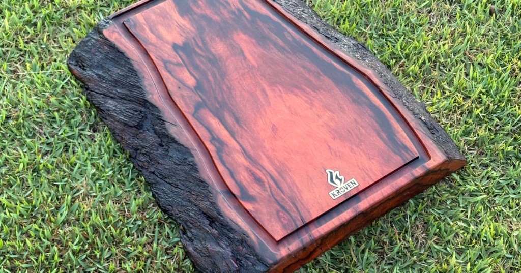 T&aacute;bua de churrasco em peroba rosa com bordas naturais da madeira, veios marcados em tons avermelhados e acabamento liso, posicionada sobre a grama.