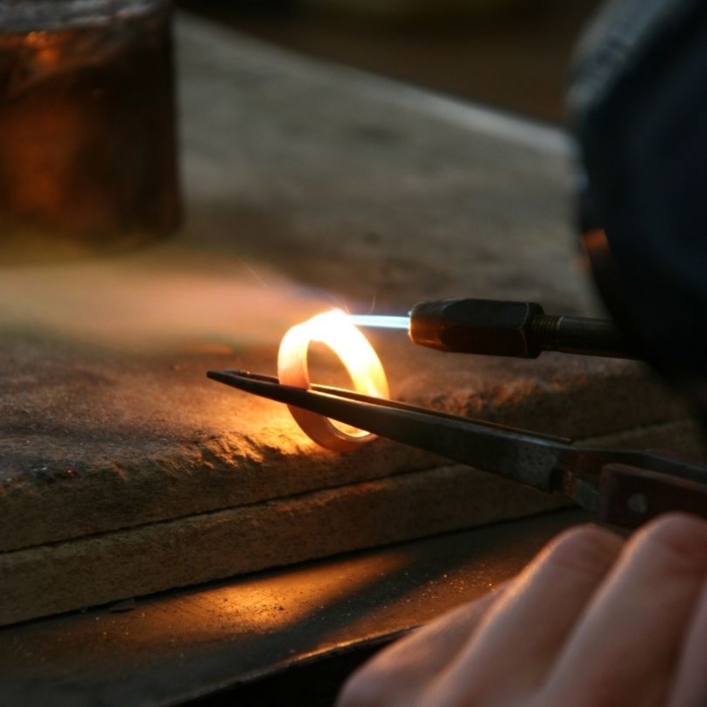 Ourives moldando uma alian&ccedil;a em moeda antiga sobre uma bancada, utilizando um ma&ccedil;arico e um pequeno alicate.