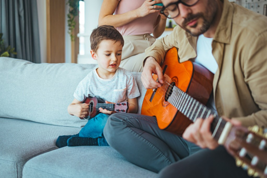 Adulto tocando viol&atilde;o enquanto crian&ccedil;a aprende m&uacute;sica com ukulele em ambiente familiar