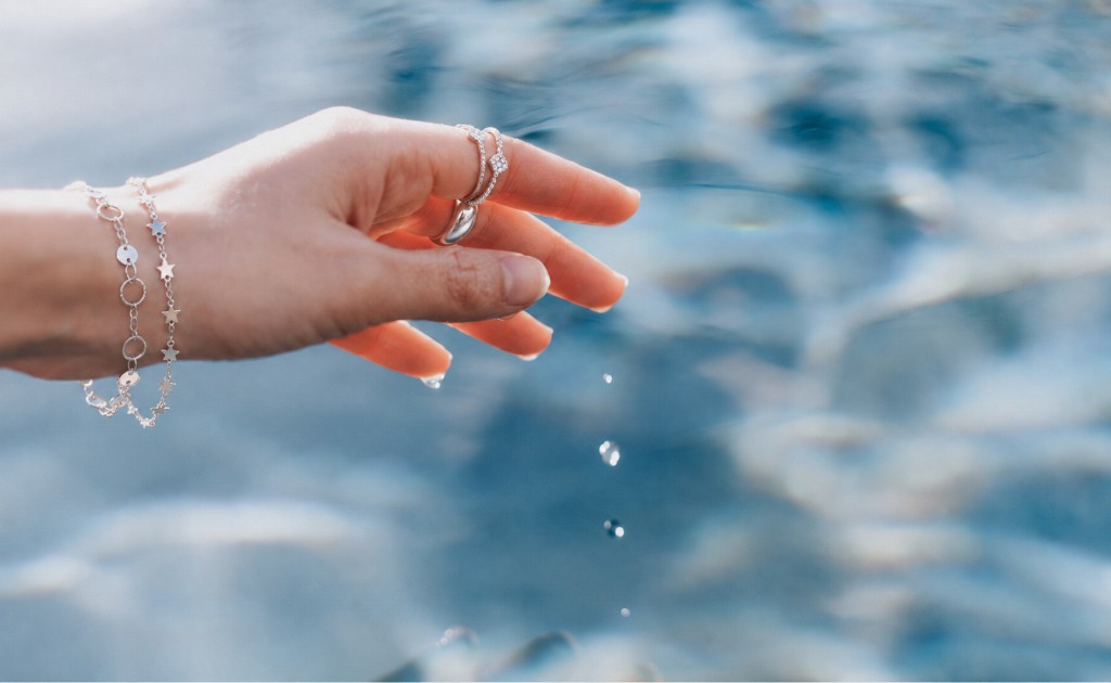 Uma m&atilde;o feminina logo acima da agua, com an&eacute;is e pulseiras prateados