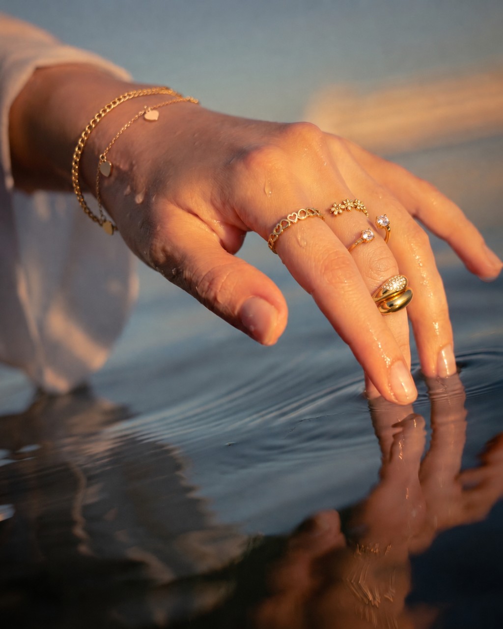 Uma mão feminina com aneis cravejados por microzircônias, pulseiras delicadas com pequenos pingentes em formato de coralçao, tocando fentilmente a água na luz do por do sol