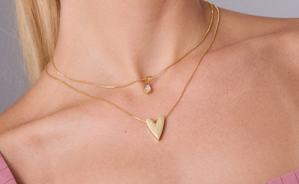Um mix de colares dourados com dois colares sobre no pesco&ccedil;o de uma modelo feminina. O colar menor tem um pingente brilhante pequeno e delicado. O segundo colar, o maior, tem um pingente em formato de cora&ccedil;&atilde;o maci&ccedil;o. Ambas as correntes no estilo persiana