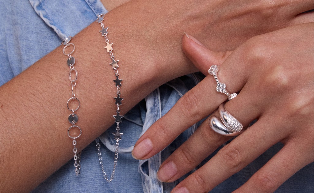 Um mix de an&eacute;is de texturas e larguras diferenres em m&atilde;os femininas ao lado de um pulso feminino com um mix de pulseiras, compondo um conjunto elegante prateado.