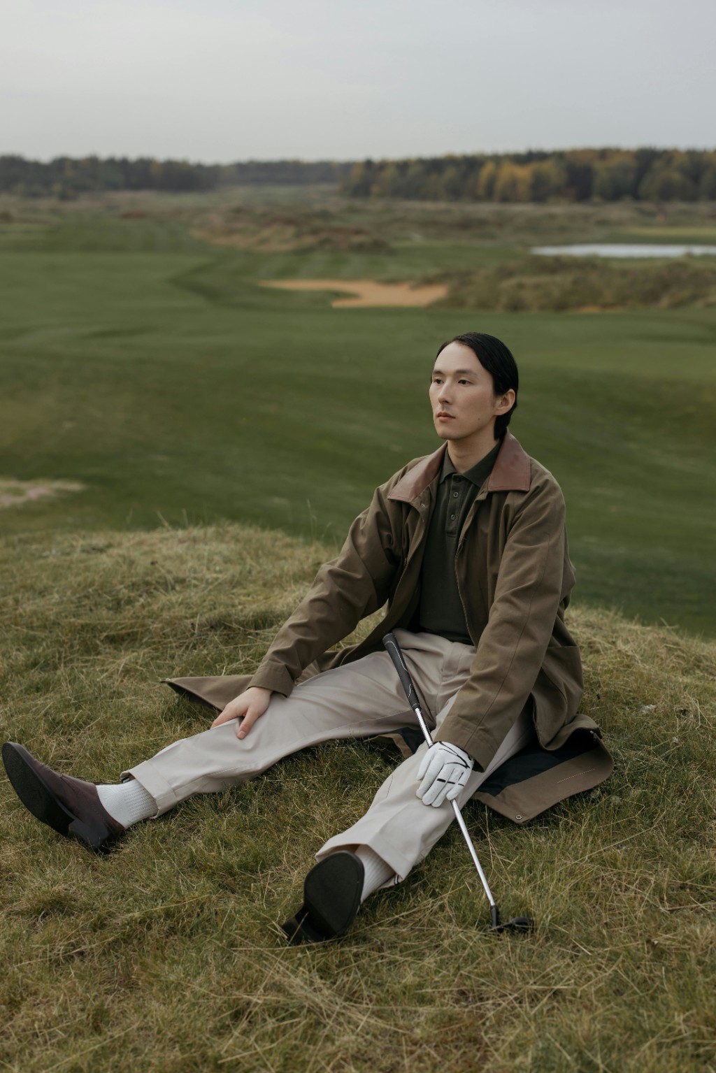 Homem asi&aacute;tico elegante sentado em campo de golfe com taco e luva contemplando o horizonte