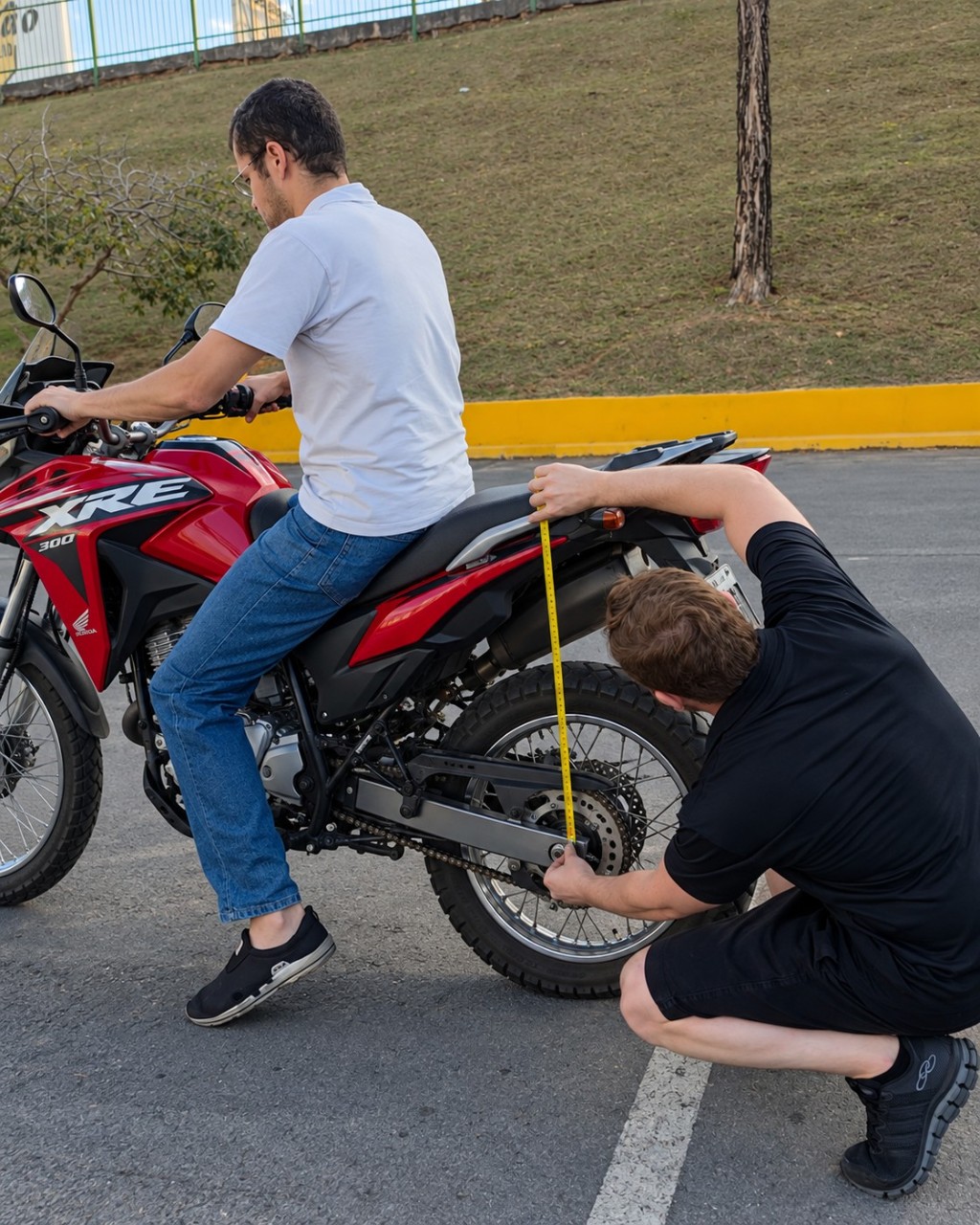 Recria&ccedil;&atilde;o por IA do apresentador GRX Parts medindo a suspens&atilde;o traseira de uma XRE 300 com o piloto sentado.