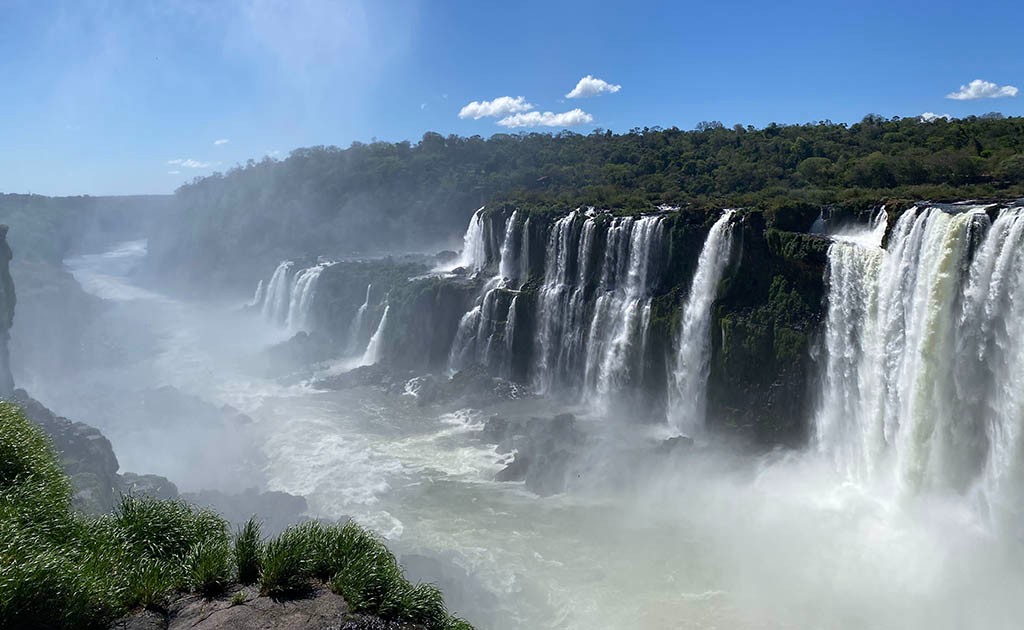 Fotograf&iacute;a panor&aacute;mica imponente de los imponentes saltos de agua en las Cataratas del Iguaz&uacute;, Misiones, Argentina, capturando la fuerza de la corriente, la densa bruma blanca y la exuberante vegetaci&oacute;n de la selva misionera bajo un cielo azul despejado.