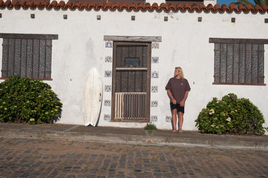 Chuck en una calle empedrada de La Pedrera junto a su tabla de surf
